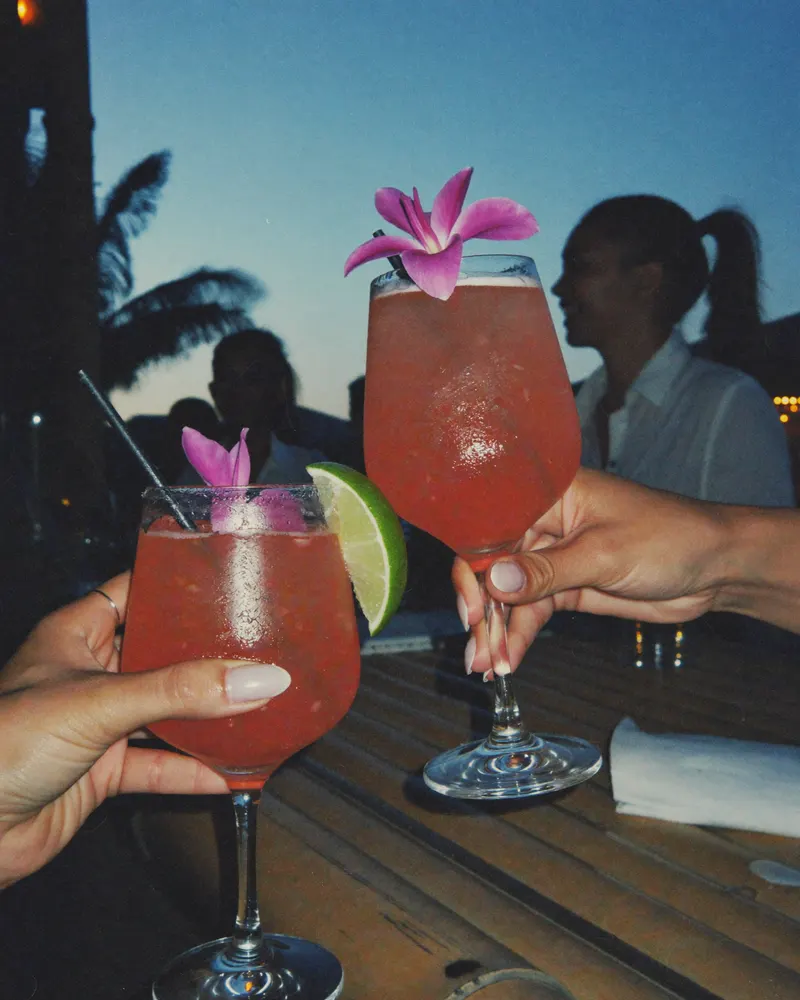 AI-generated image: friends toasting at a bar at dusk, frangipani on the table